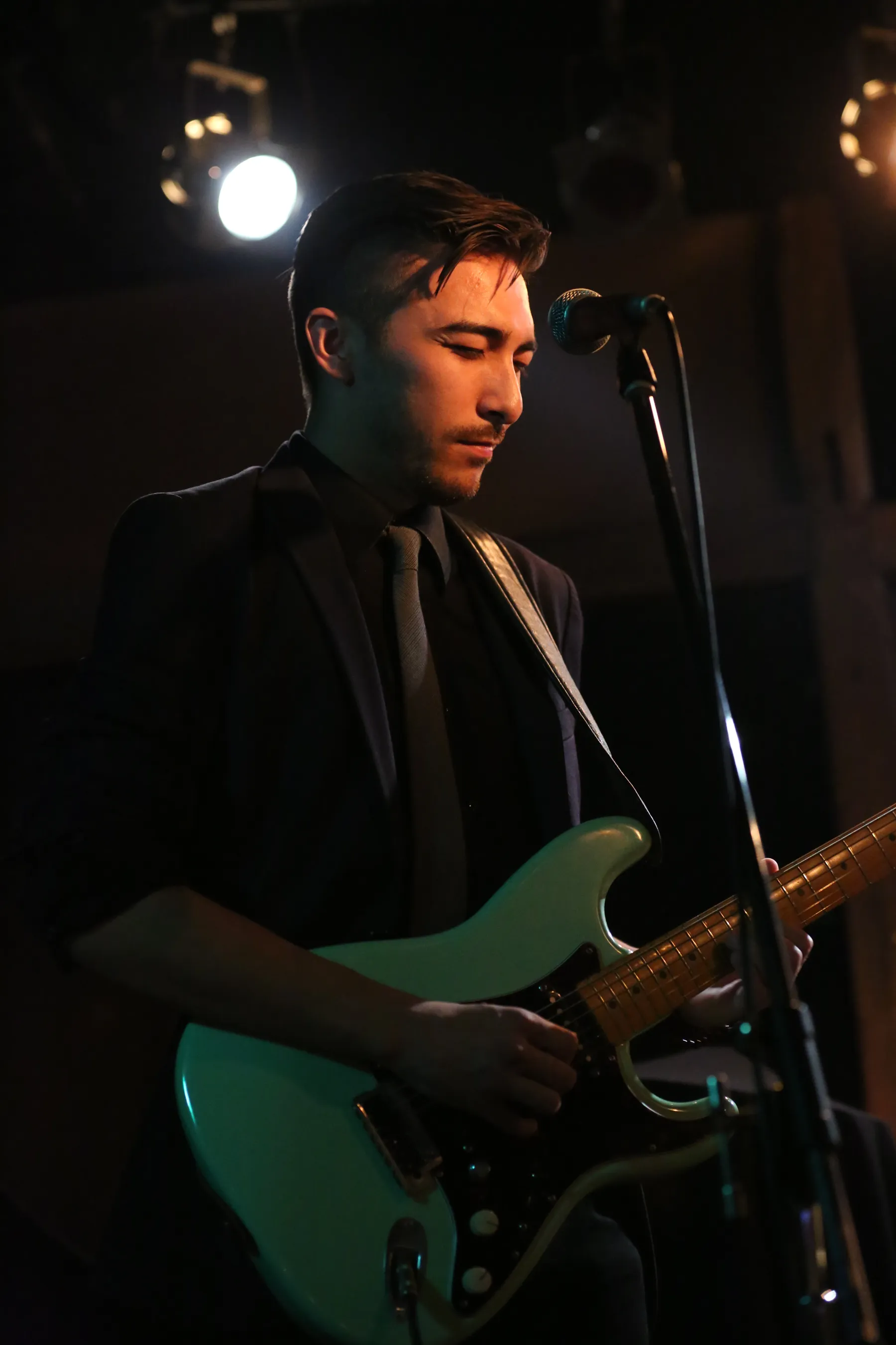 Alex Nishi on stage in a suit, playing a Stratocaster under stage lights